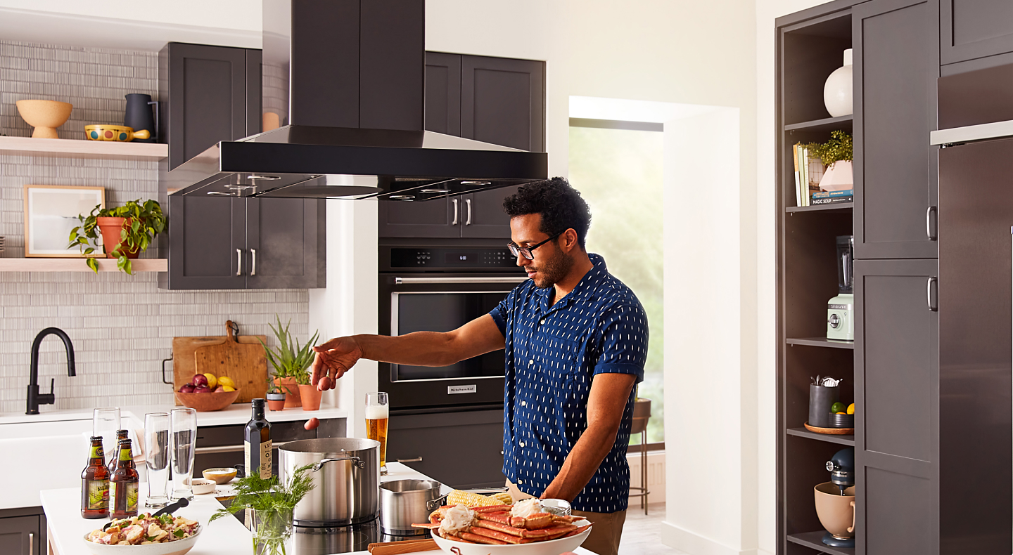 A person adding food to a pot on a KitchenAid® cooktop with a range hood above