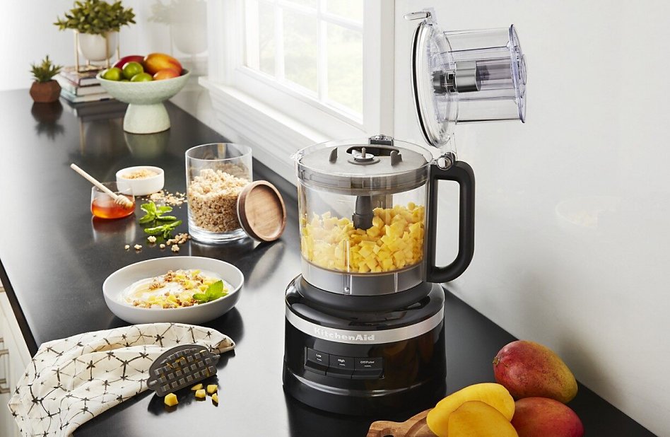 Large-sized black food processor with diced mango on counter with other ingredients Large-sized black food processor with diced mango on counter with other ingredients