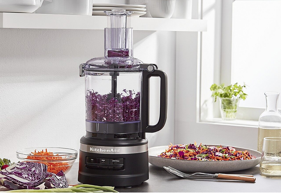 Black food processor with shredded red cabbage on counter with salad
