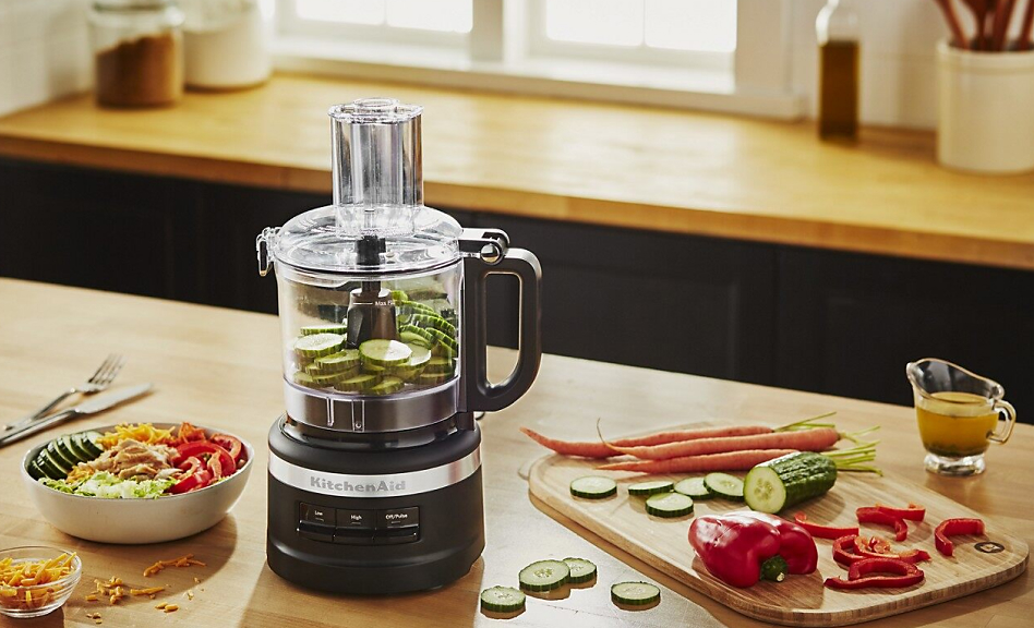 Black food processor with sliced cucumbers surrounded by vegetables and a salad