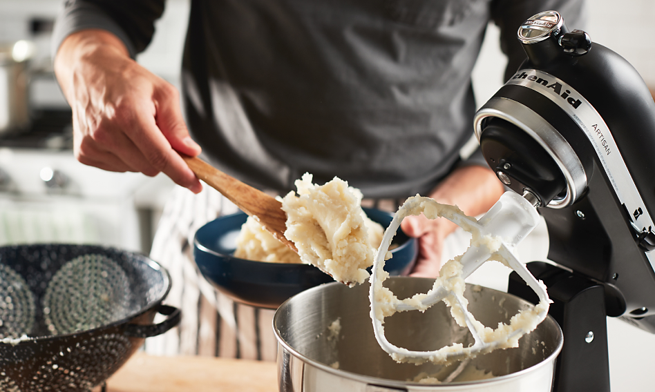 Male hand holding a bowl and a wooden spoon filled with mashed potatoes over a KitchenAid brand stand mixer