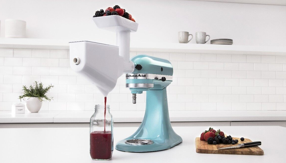 Blue KitchenAid brand stand mixer with berries inside juicing attachment and a cutting board with berries on the countertop
