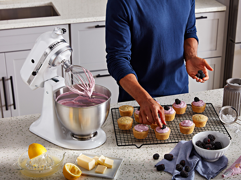 White KitchenAid® stand mixer next to person putting toppings on cupcakes