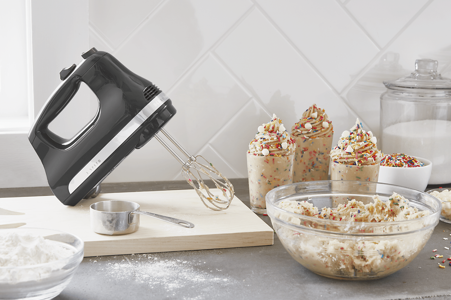 Black KitchenAid® hand mixer on wooden cutting board next to whipped cream with sprinkles in serving cups