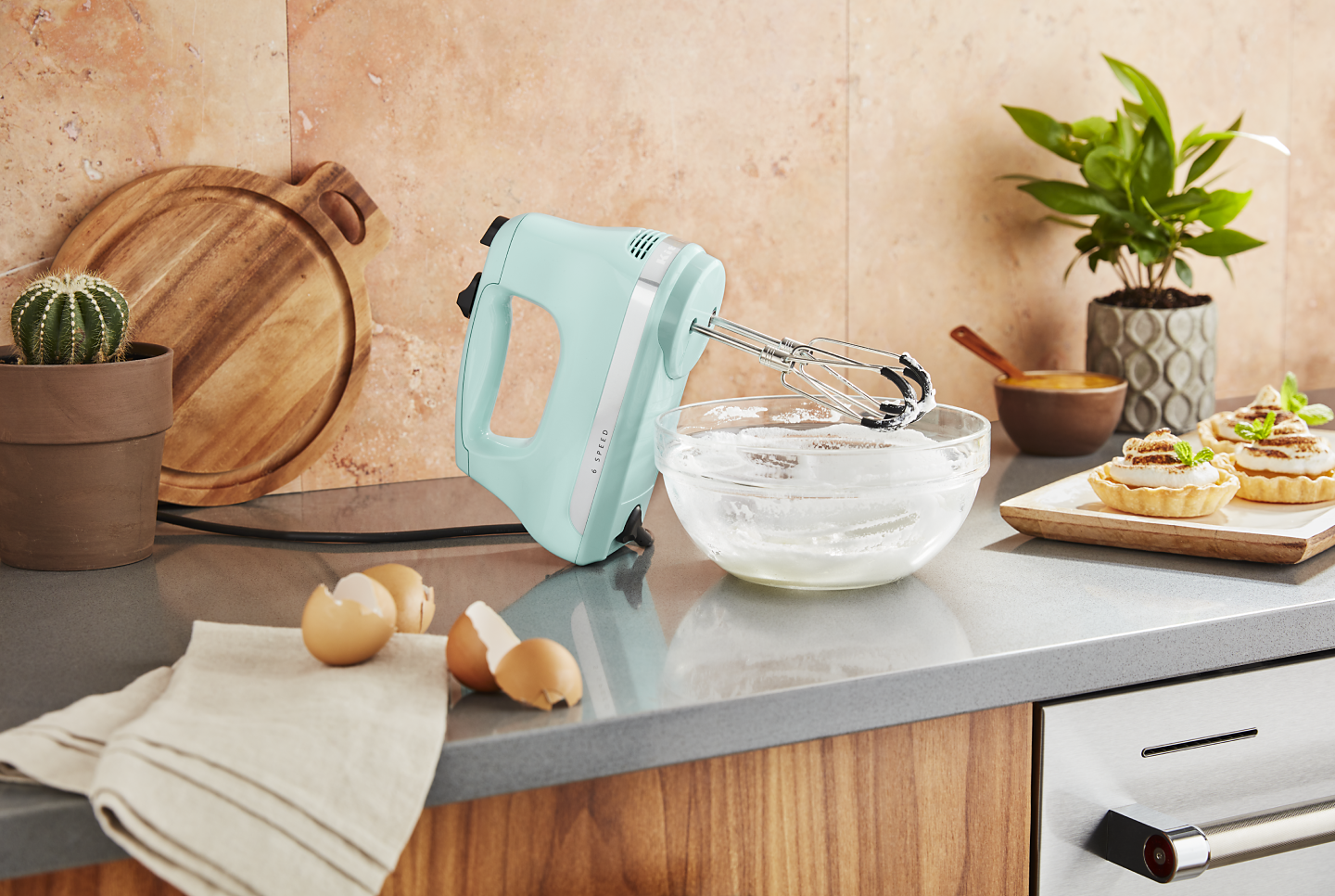 Light pistachio green KitchenAid® hand mixer on countertop with egg shells and bowl of whisked egg whites