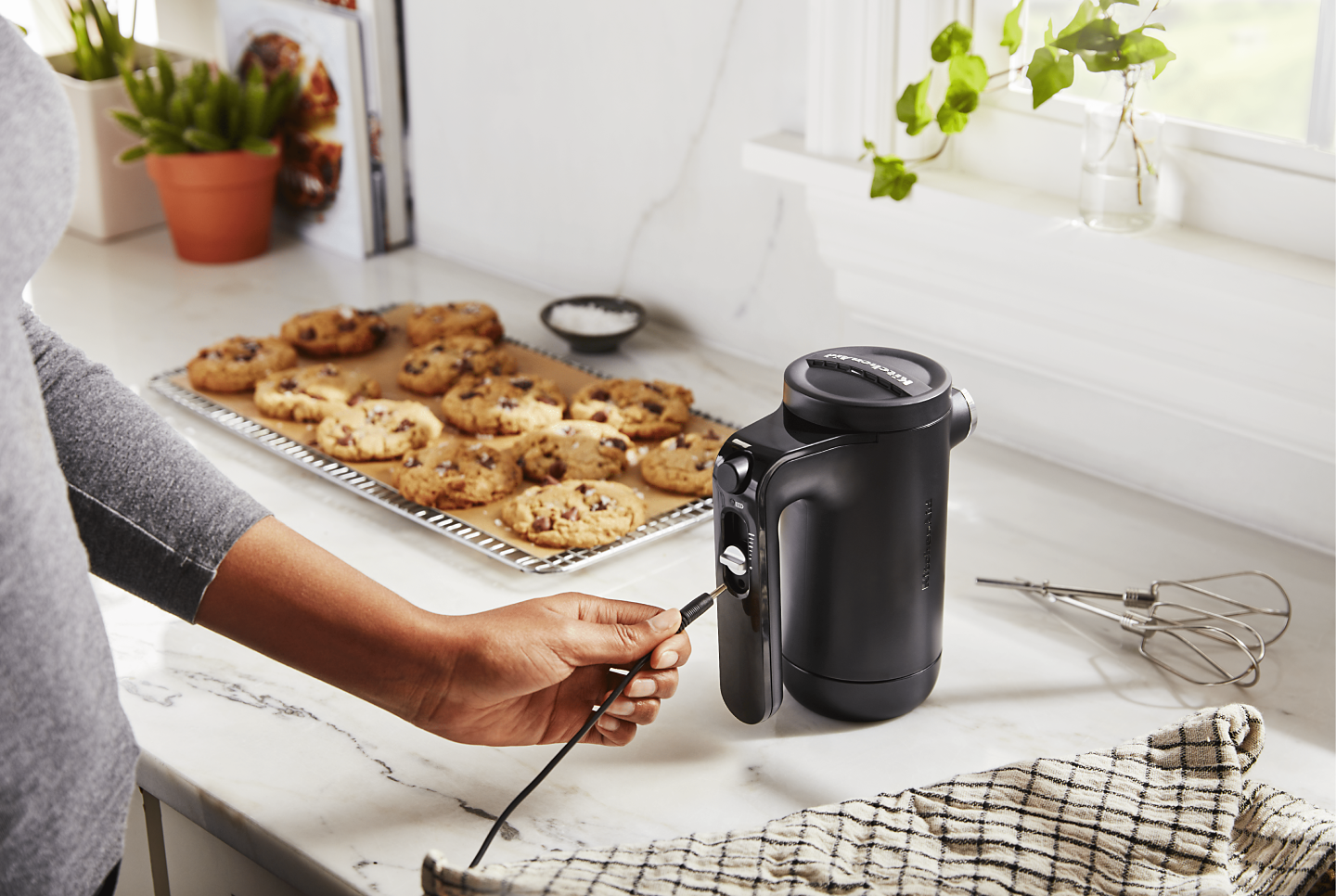 Maker plugging cord into black KitchenAid® hand mixer on countertop next to tray of chocolate chip cookies