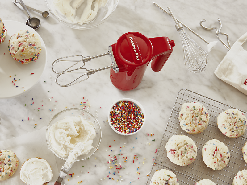 Red KitchenAid® hand mixer next to bowl of sprinkles, icing and bath of homemade cookie cream pies