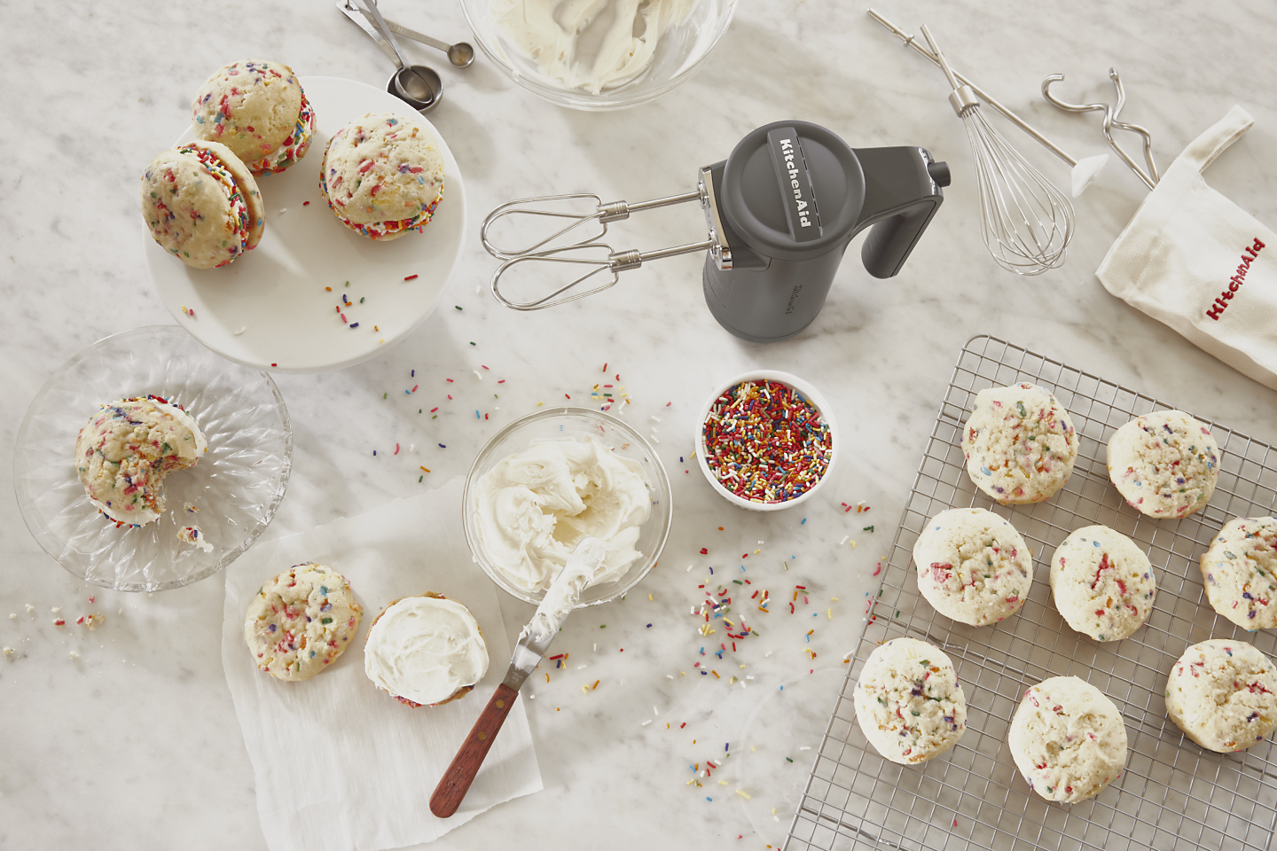 Gray KitchenAid® hand mixer standing upright on countertop along with sandwich cookies, frosting, sprinkles and beater accessories