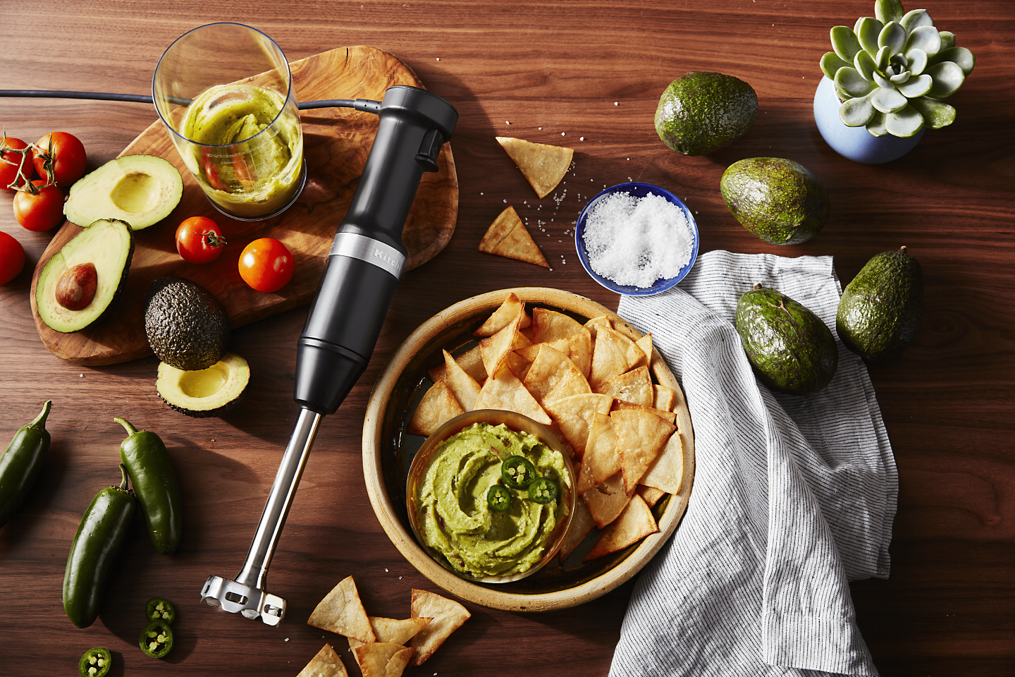 Black KitchenAid® immersion blender on wood surface with guacamole ingredients and blended guacamole with chips