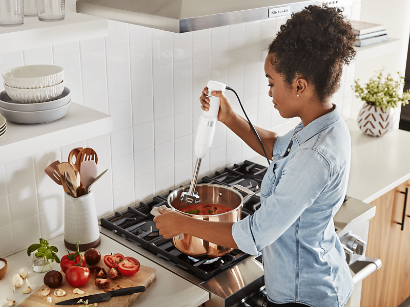 Maker using KitchenAid® immersion blender to mix ingredients in pot on top of stovetop