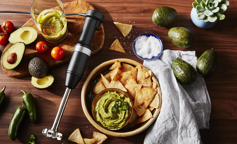  Immersion blender laying beside bowl of guacamole and chips surrounded by guacamole ingredients