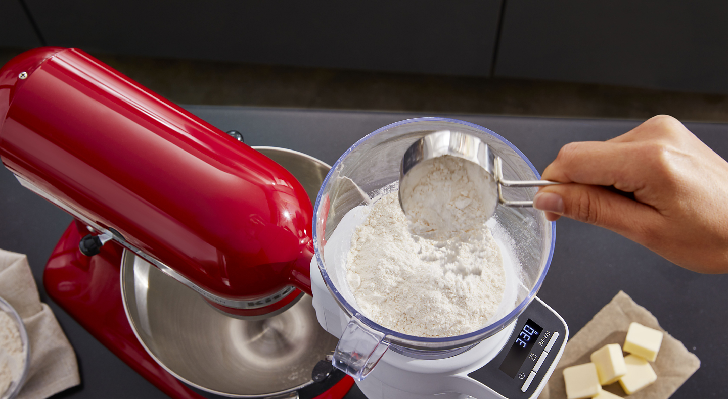 A person sifting flour into a red KitchenAid® stand mixer.