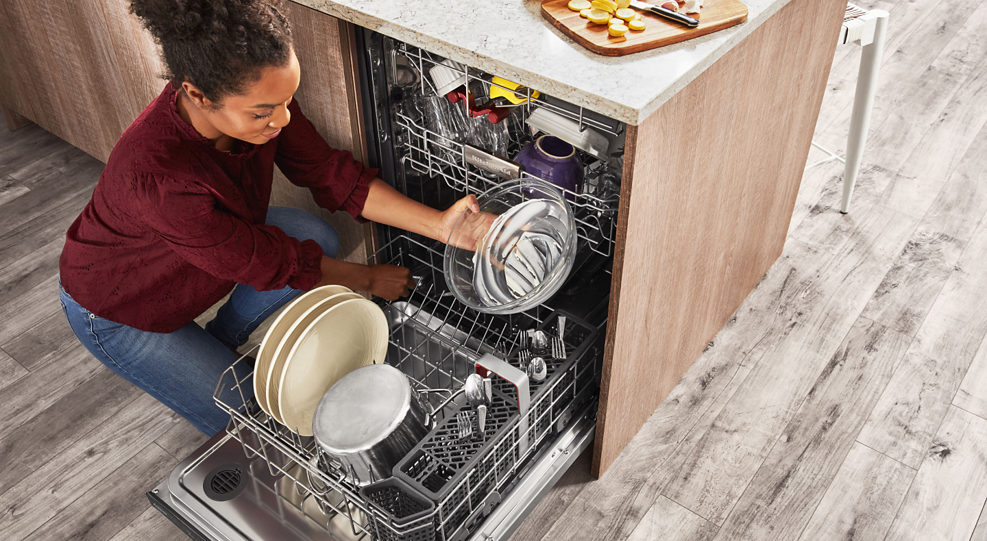 A person removing clean dishes from the bottom rack of a KitchenAid® dishwasher.