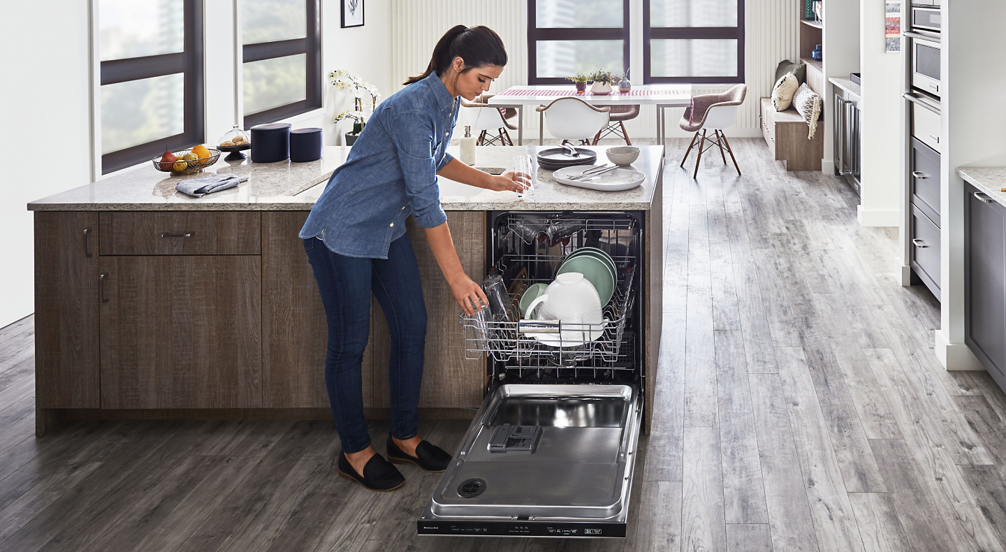 A person placing a glass on the top rack of a KitchenAid® dishwasher in a modern kitchen.