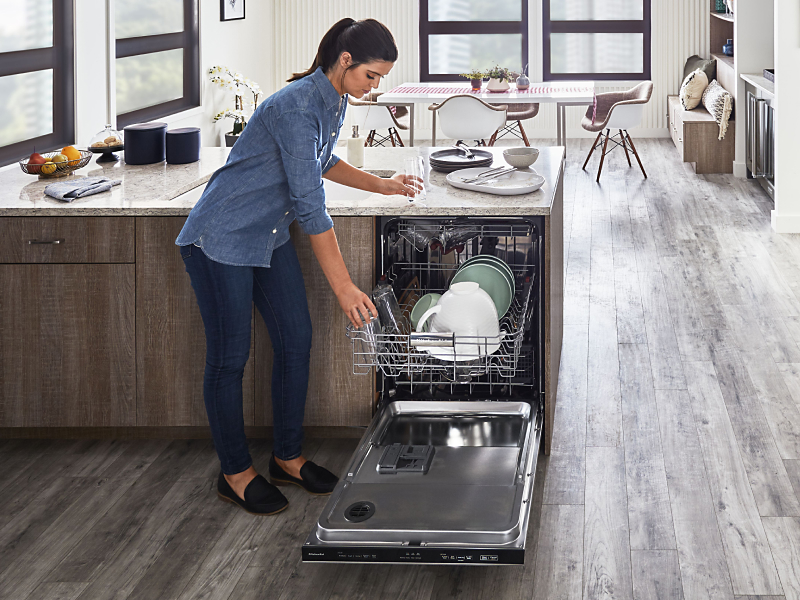 A person placing a glass on the top rack of a KitchenAid® dishwasher in a modern kitchen.