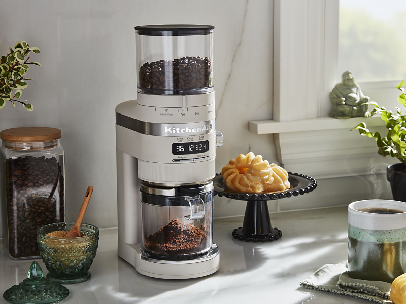 A KitchenAid® burr coffee grinder in a modern kitchen next to a pastry and cup of coffee.