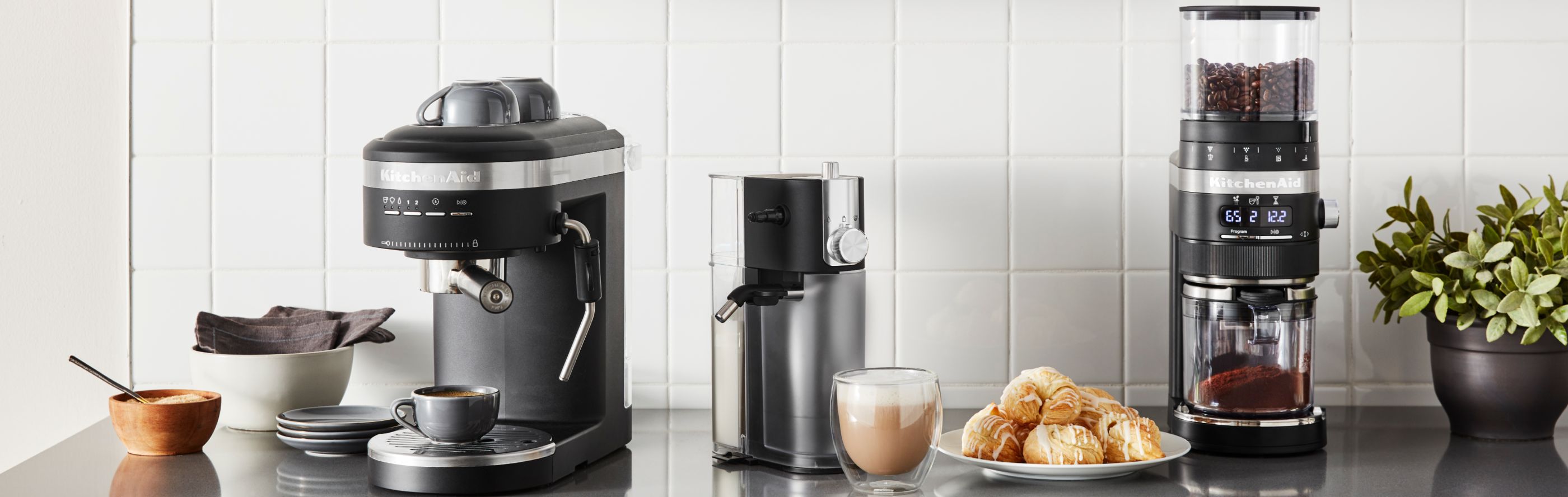 A KitchenAid® espresso machine and burr coffee grinder in a modern kitchen next to a frothy cappuccino and pastries.