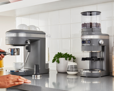 A woman using a KitchenAid® espresso machine next burr coffee grinder in a modern kitchen