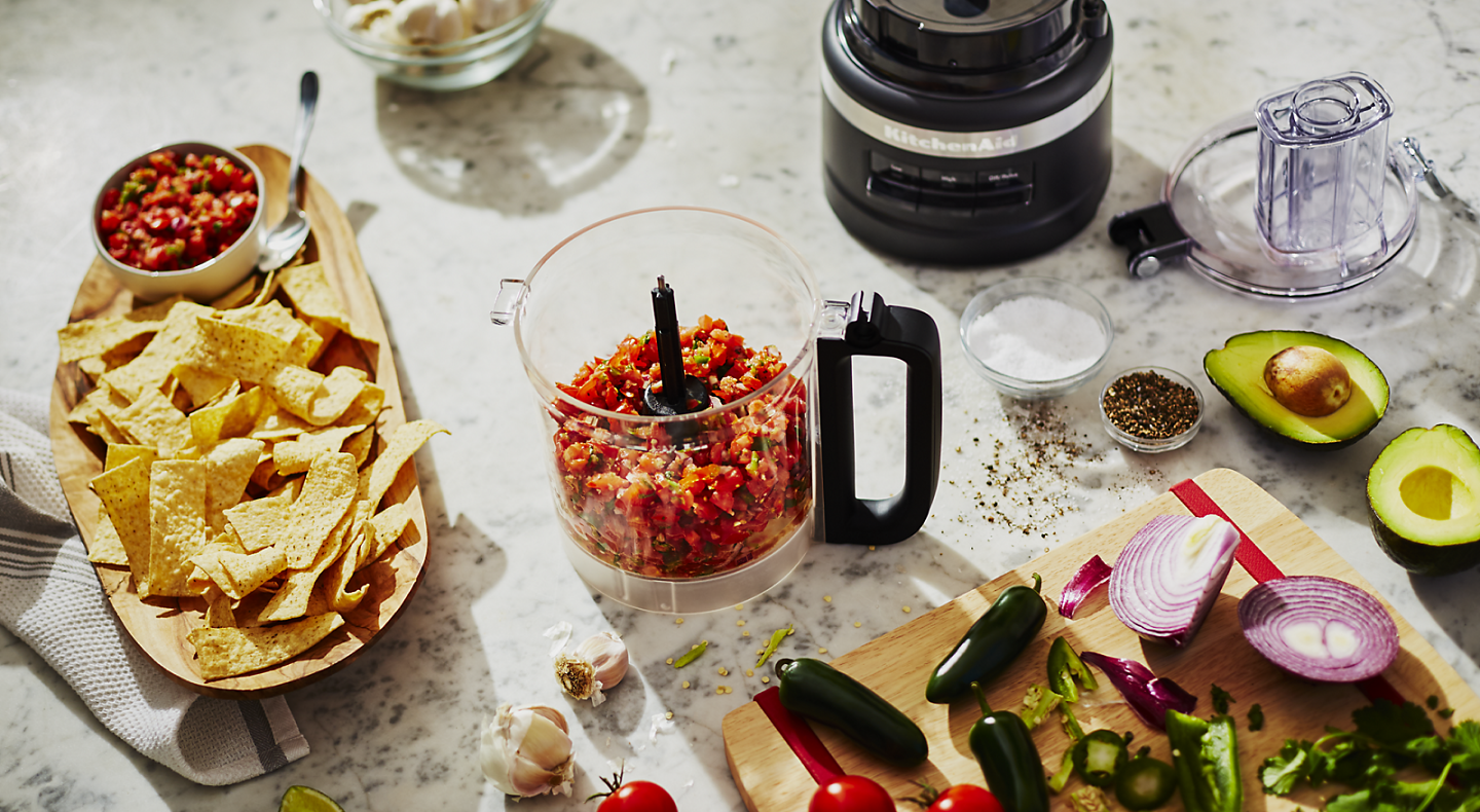 A food processor filled with salsa surrounded by the ingredients used in its creation.