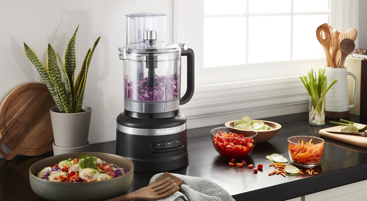 A black table that holds a food processor chopping onions as well as a fresh salad.