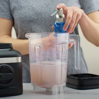 Person dispensing dish soap into KitchenAid® blender jar half-filled with water next to black base