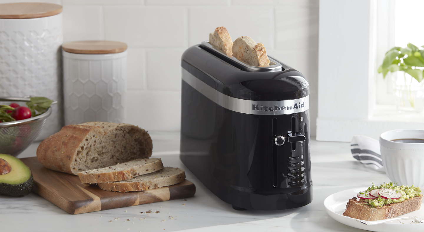 Slices of bread in a black KitchenAid® toaster next to a loaf of artisan bread and an open-faced sandwich on a modern kitchen counter.