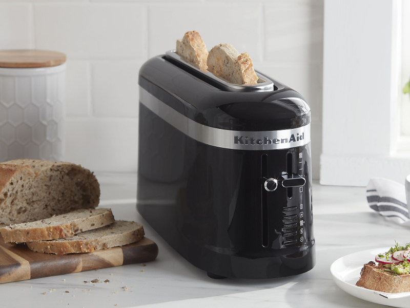 Slices of bread in a black KitchenAid® toaster next to a loaf of artisan bread and an open-faced sandwich on a modern kitchen counter.