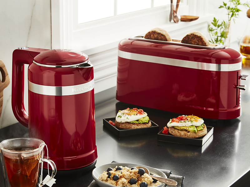 Slices of bread in a red KitchenAid® toaster next to open-faced sandwiches and cups of tea on a modern kitchen counter.