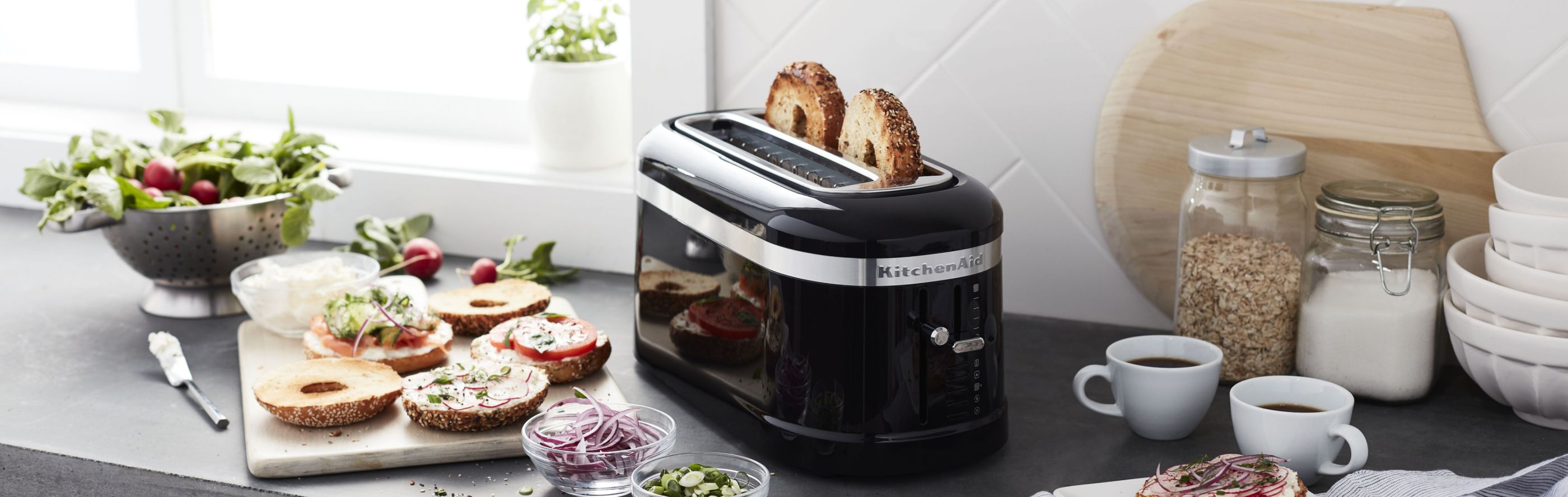 Bagel slices in a pop-up KitchenAid® toaster on a modern kitchen counter next to bagel sandwich fixings.