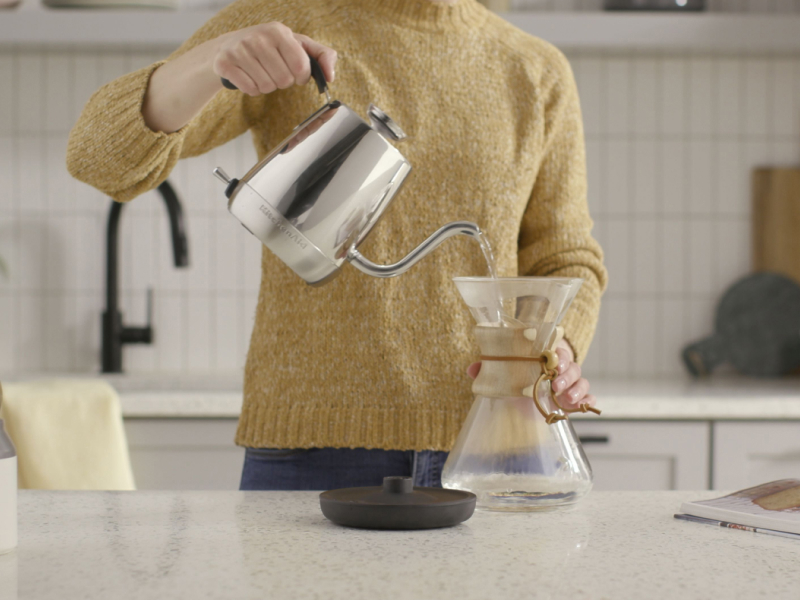 A woman in a modern kitchen pouring water from a KitchenAid® electric kettle into a glass container