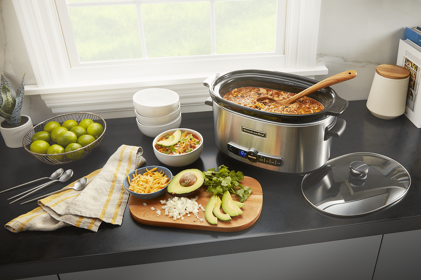KitchenAid® slow cooker filled with chili next to a cutting board of vegetables and herbs