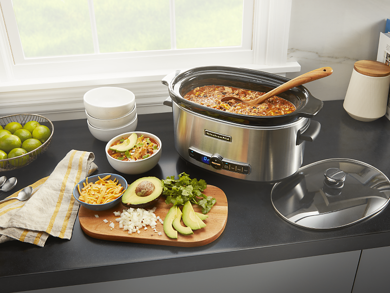 KitchenAid® slow cooker filled with chili next to a cutting board of vegetables and herbs