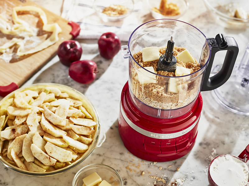 Crushed graham crackers and butter inside a KitchenAid® food processor next to an apple pie
