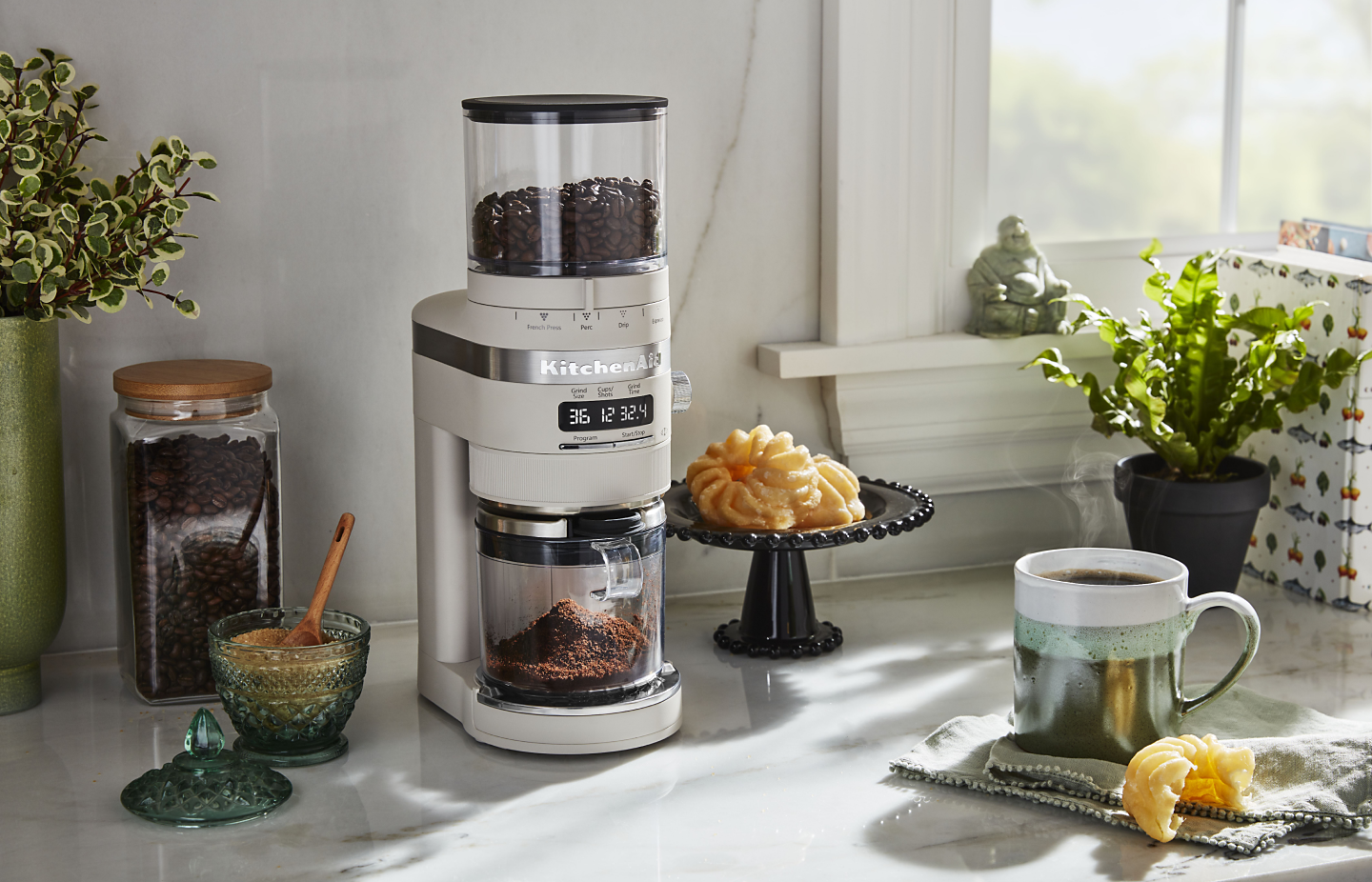 White KitchenAid® burr grinder next to a cup of coffee and pastries