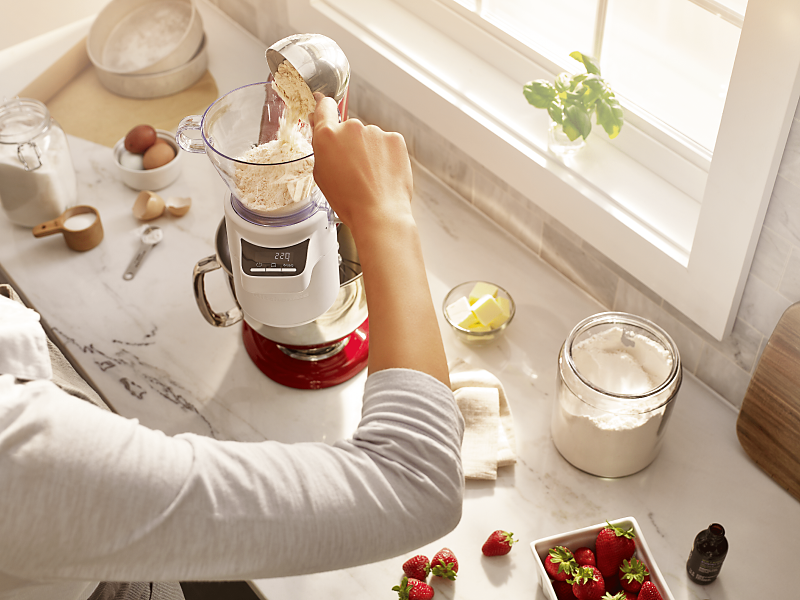 A woman adding flour to the KitchenAid®  stand mixer sifter + scale attachment. 