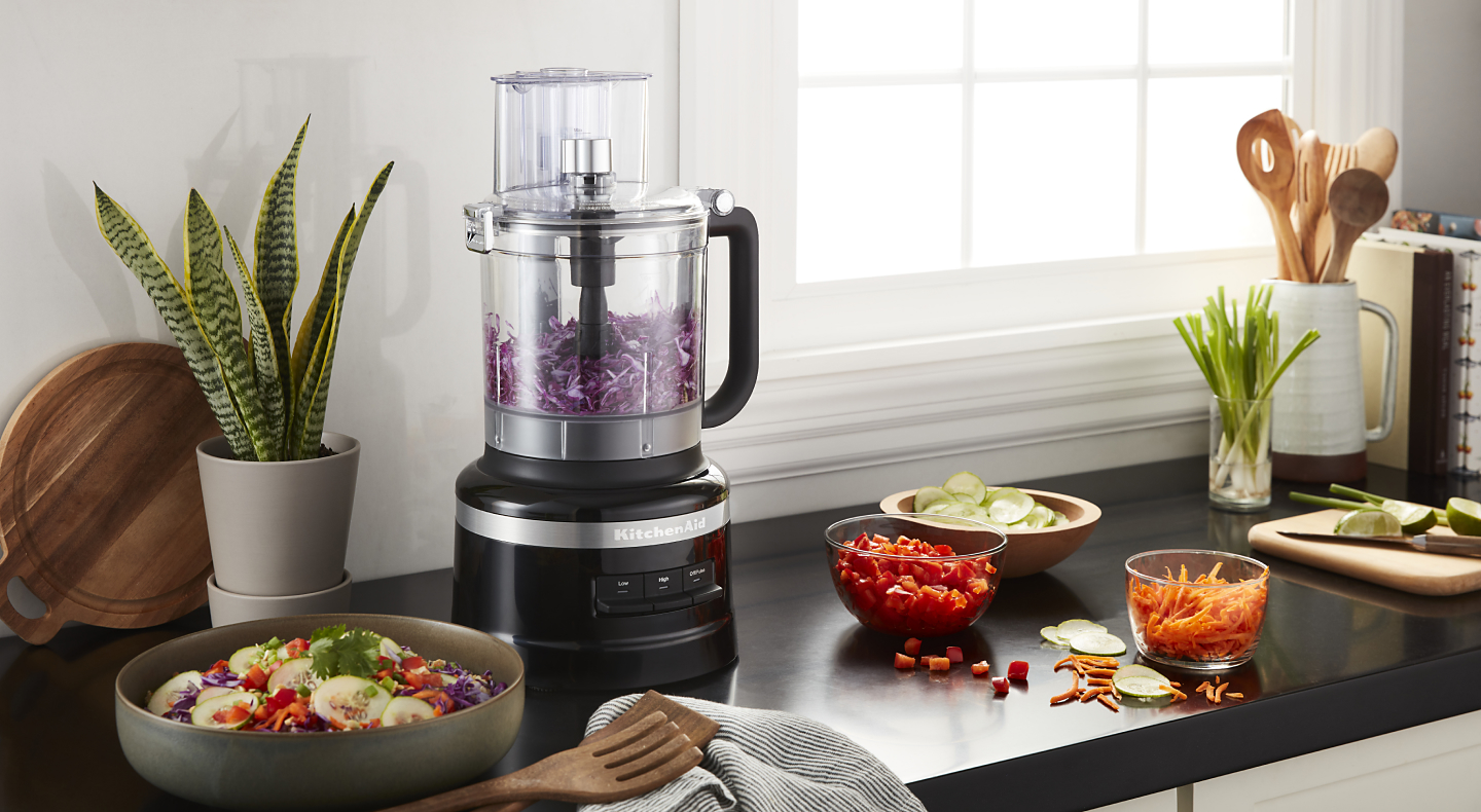 KitchenAid® food processor with purple cabbage next to a chopped salad and the ingredients in a modern kitchen.