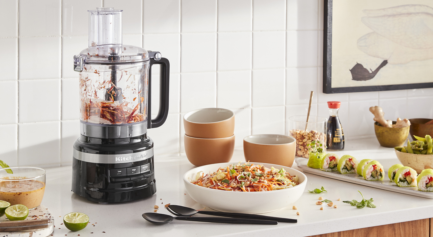 KitchenAid® food processor with chopped vegetables next to a chopped salad and freshly made salad dressing in a modern kitchen.