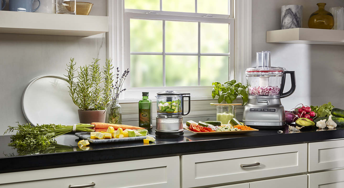 KitchenAid® food processor and food chopper with chopped vegetables next to fresh vegetables in a modern kitchen.