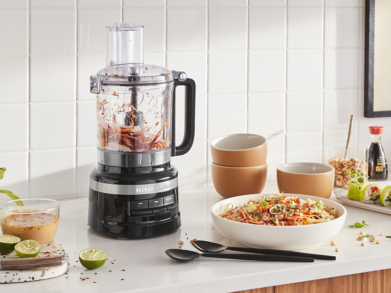 KitchenAid® food processor with chopped vegetables next to a chopped salad and freshly made salad dressing in a modern kitchen.