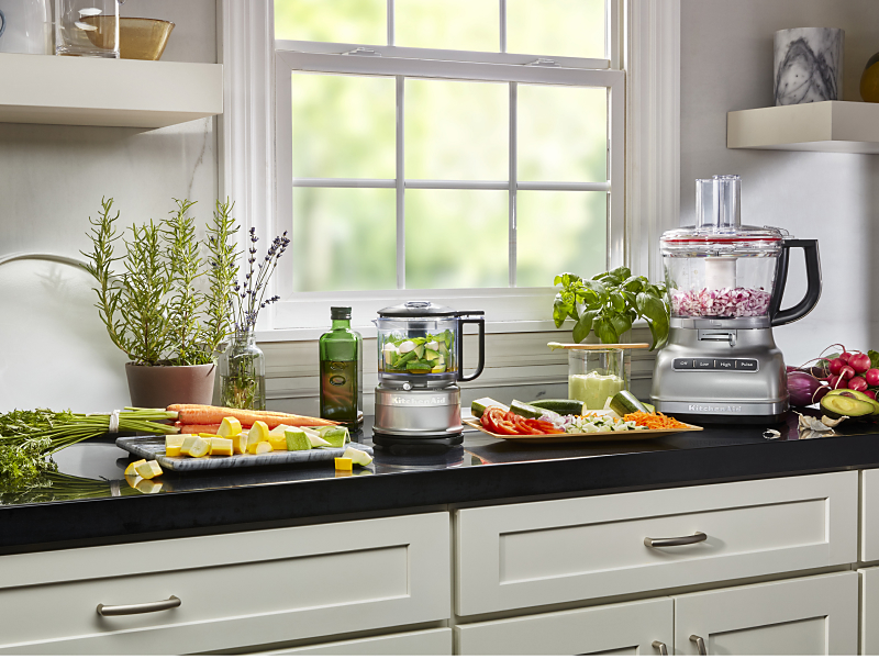 KitchenAid® food processor and food chopper with chopped vegetables next to fresh vegetables in a modern kitchen.
