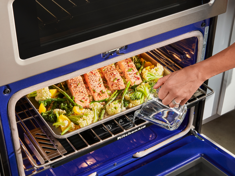 A woman checking on salmon and vegetables on a baking sheet in a KitchenAid® oven.
