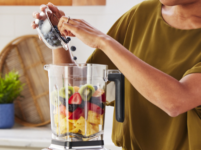 A woman adding blueberries to a KitchenAid® blender that has a variety of fruit