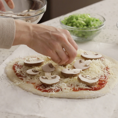 Person adding sliced mushrooms on top of a homemade pizza