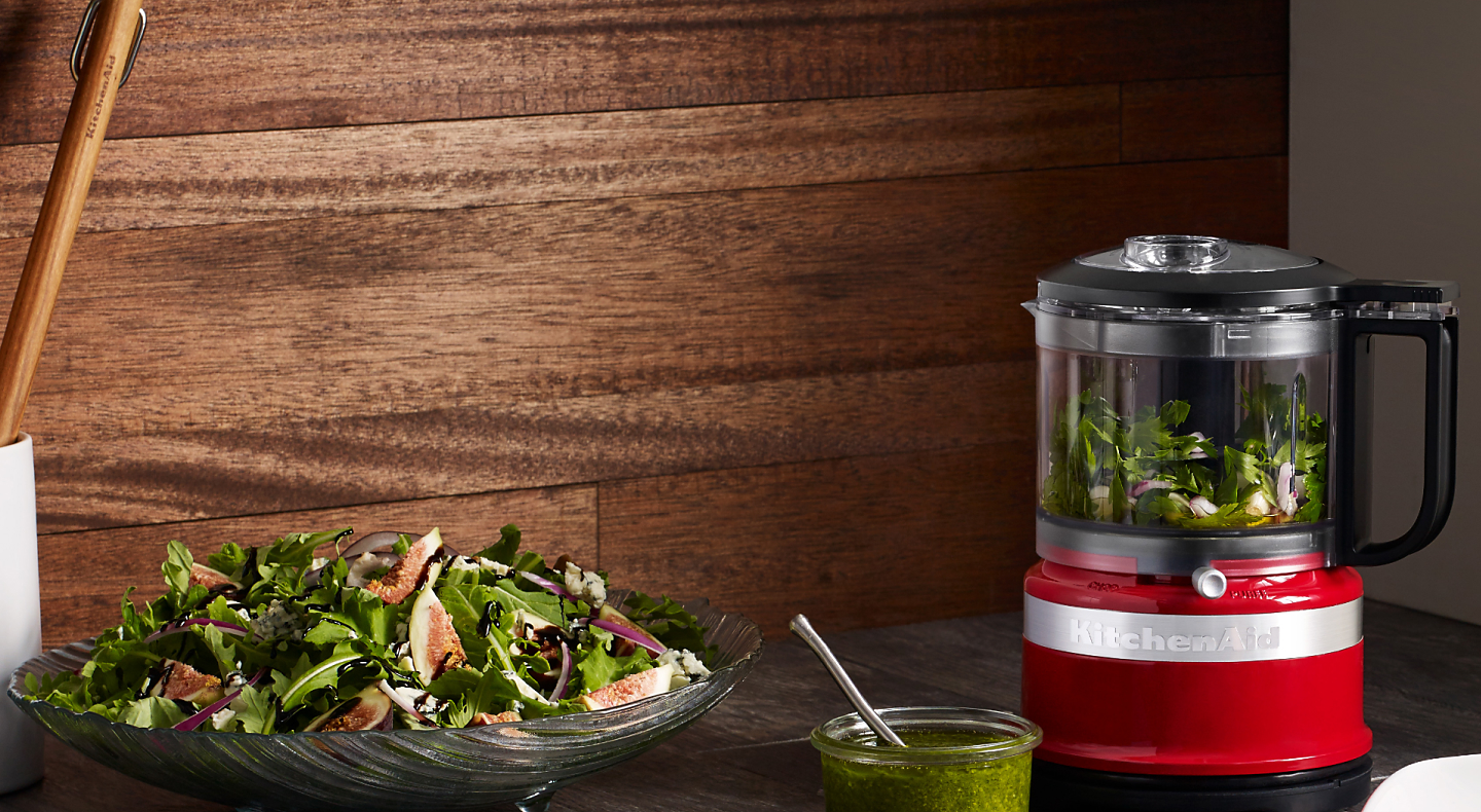 Small red food processor filled with mixed greens next to tossed salad on counter