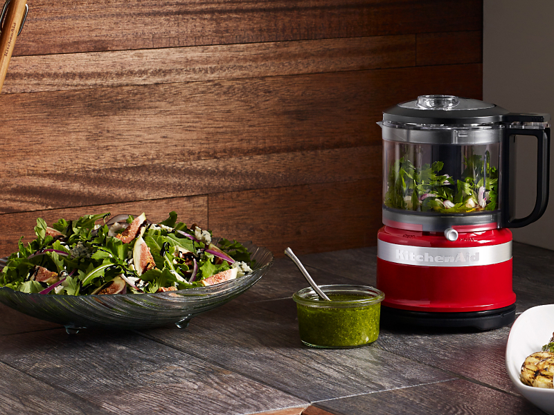 Small red food processor filled with mixed greens next to tossed salad on counter