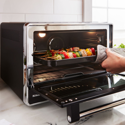 A person holding a tray of kebabs cooking inside a black countertop oven