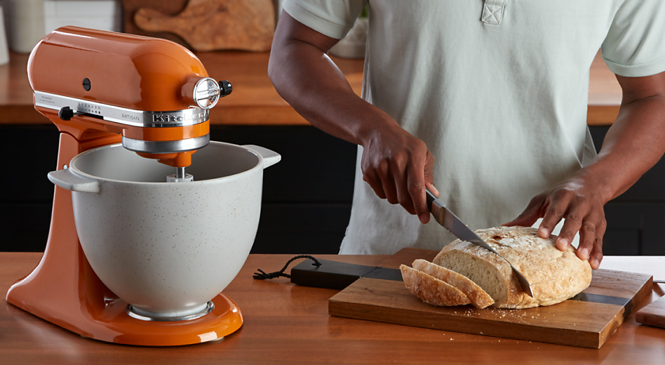 An orange stand mixer and a person slicing homemade bread.