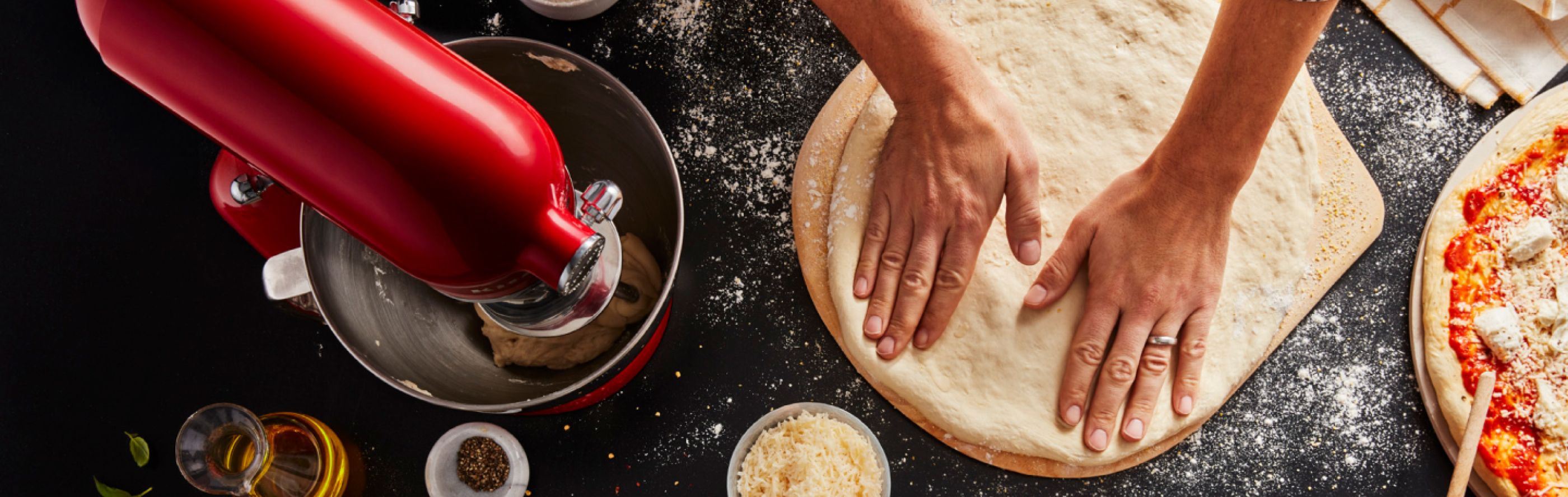 Maker flattening out pizza dough on countertop with KitchenAid® stand mixer and pizza ingredients