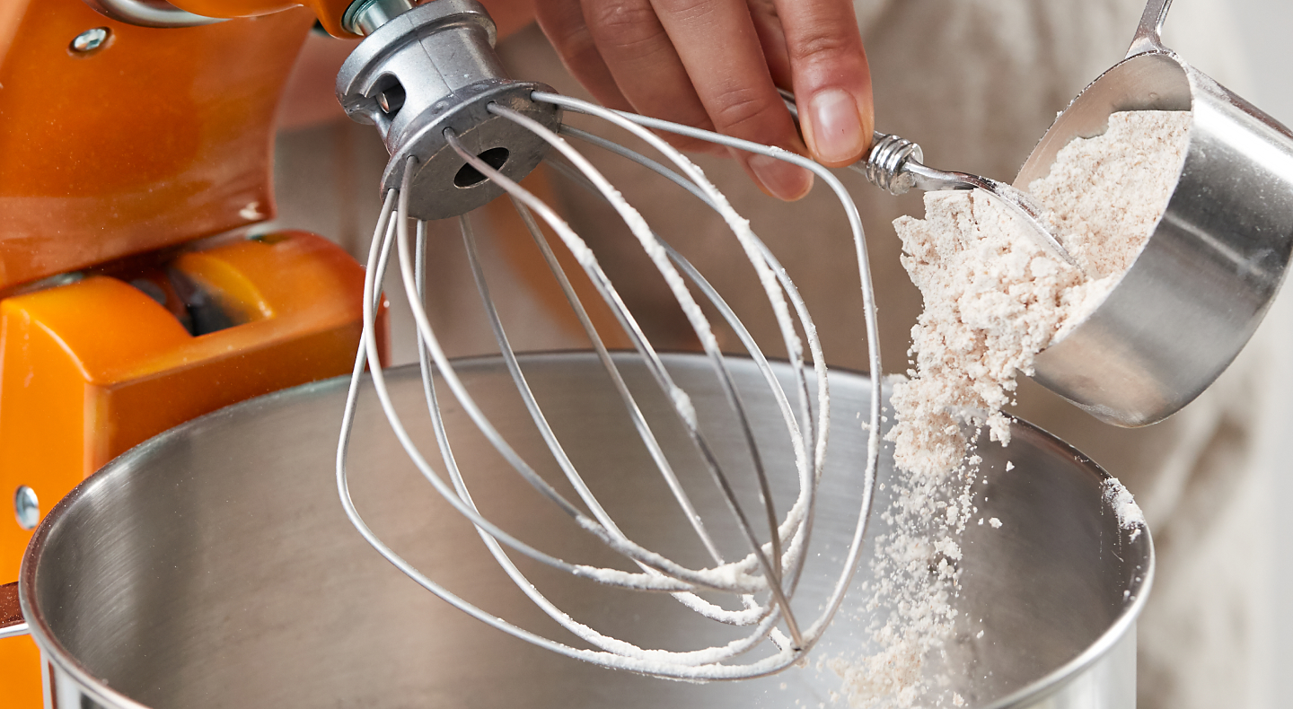 Person adding flour to stand mixer bowl beneath whip attachment