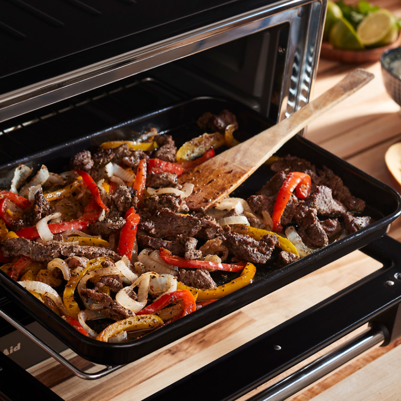 Meat and vegetables in tray of open air fryer with wooden spatula.